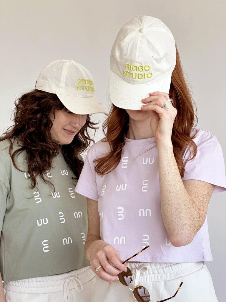 A woman in pistachio crop tee and white Ringo Studio cap, posing next to studio founder wearing Ringo Studio lavender tee and Ringo Studio white cap.