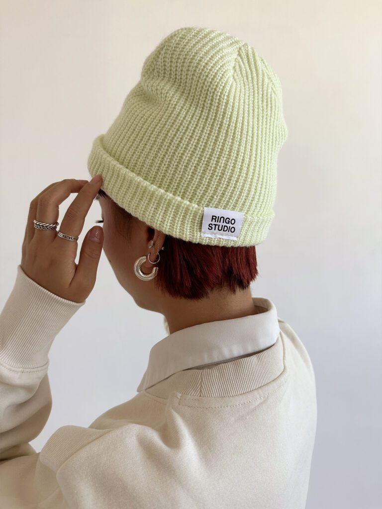 Woman wearing beige Puff print sweatshirt with the lime Ringo Studio beanie, with the Ringo Studio label tag peeking.